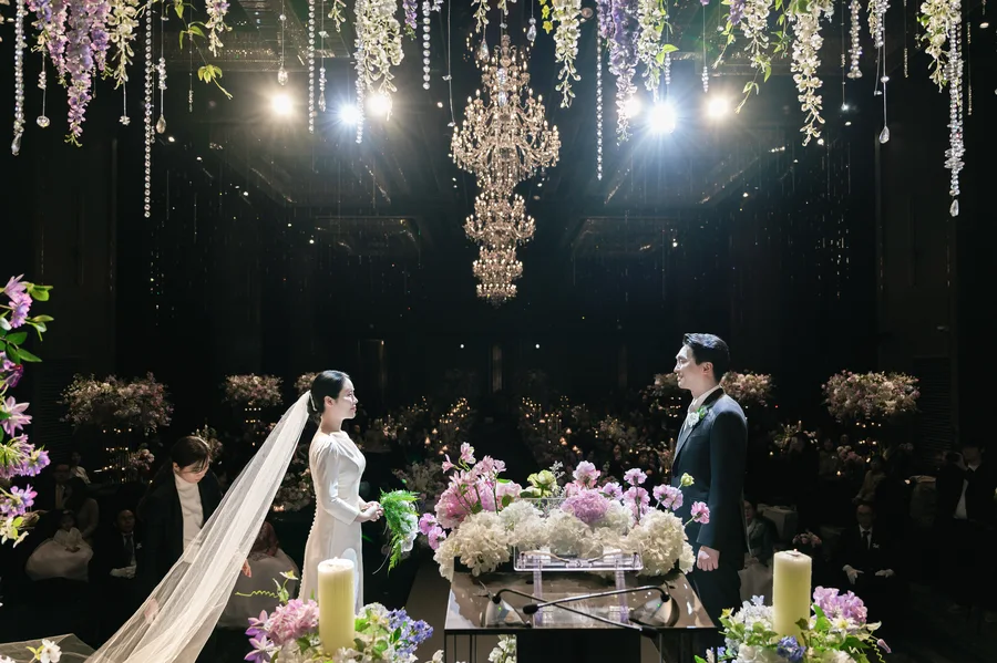 늘어진 플라워 샹들리에 아래 주례상 사이에 마주 선 신랑신부