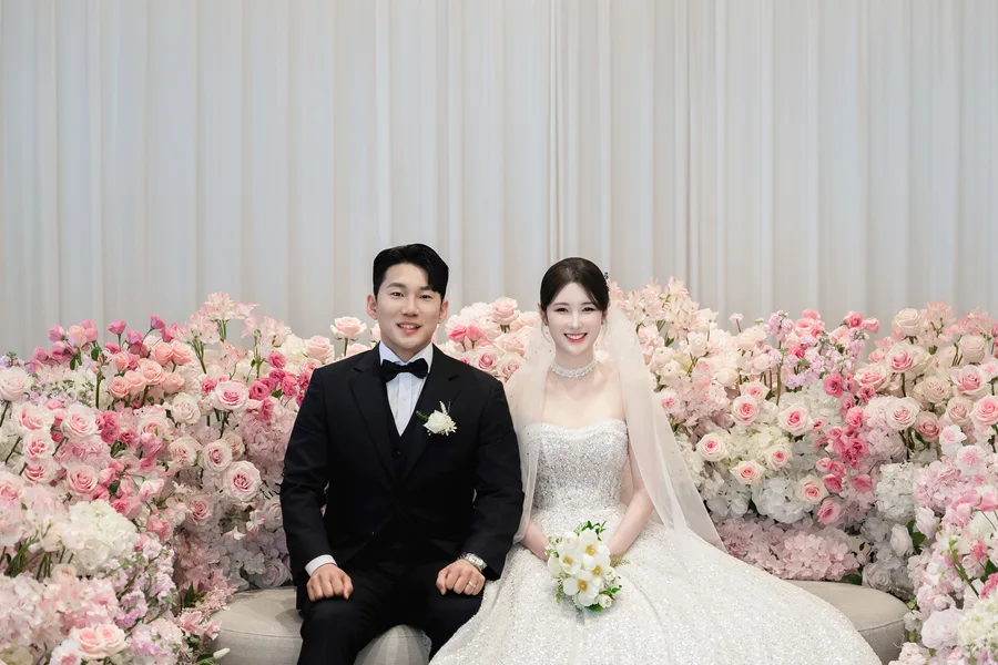 분홍 꽃벽 앞 의자에 나란히 앉아 카메라를 보고 웃는 신랑신부