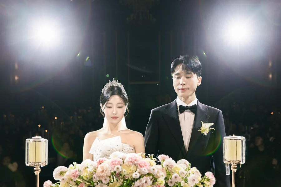 촛대와 꽃 장식을 사이에 두고 나란히 선 신랑신부, 강한 역광 인물컷